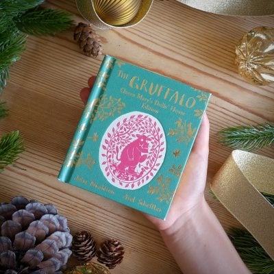 Hand holding a teal box with a pink logo on a wooden surface with decorative elements.