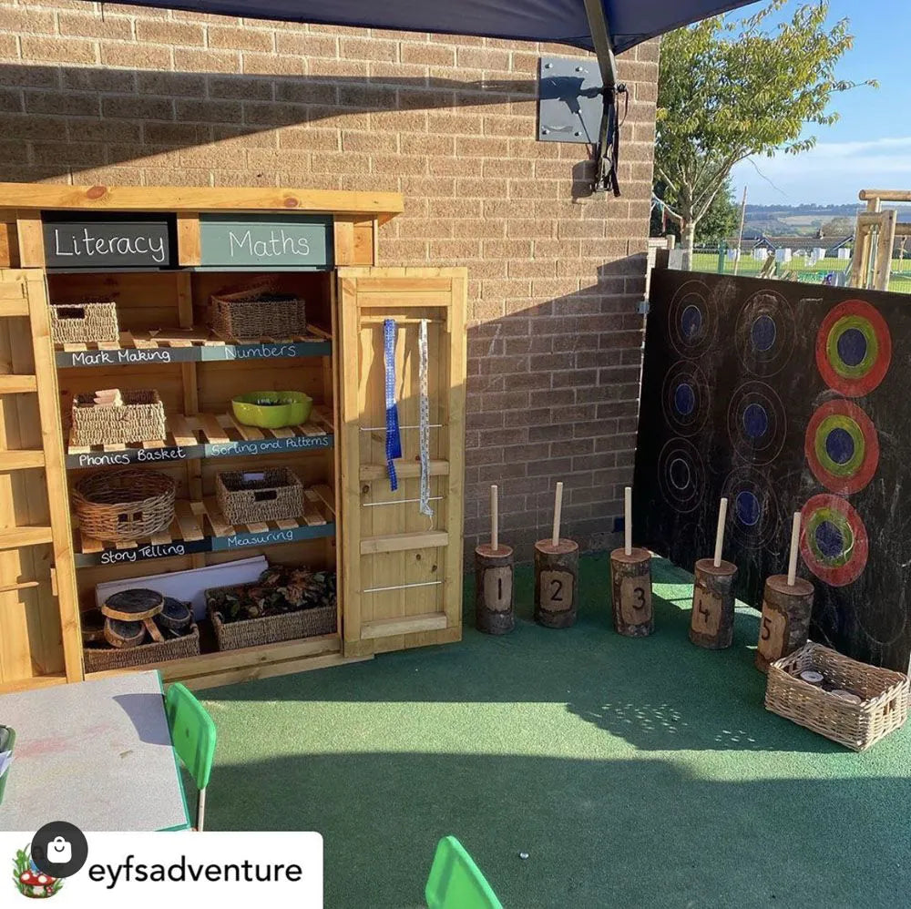 Educational outdoor play area with wooden shelves labeled 'Literacy' and 'Maths', number drums, and a target board.