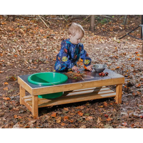 Child playing at a wooden outdoor table with green and silver bowls on a leaf-covered ground.