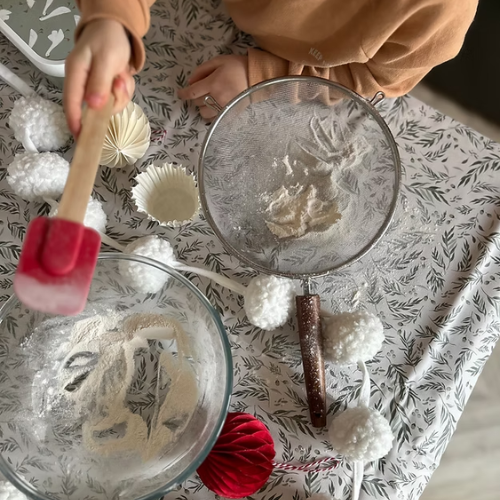 Person sifting flour into a bowl with decorative items on a patterned tablecloth.
