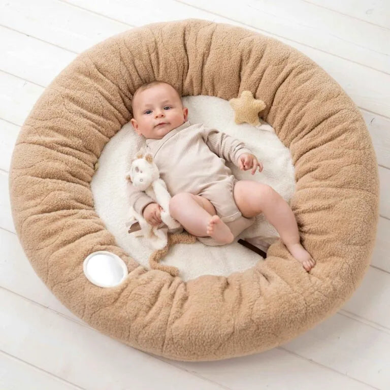 Baby lying on a round beige cushion with a white blanket, holding a plush toy on a light wooden floor.