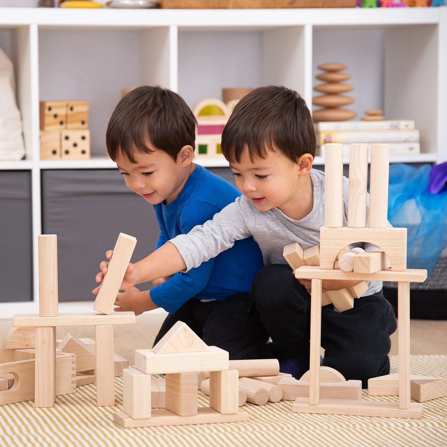 Two children playing with wooden building blocks in a room with shelves in the background.
