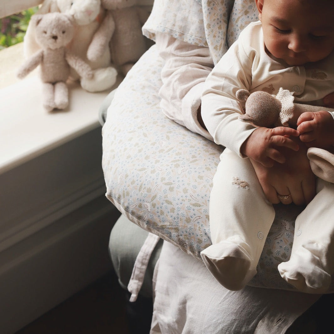 Baby in white outfit holding a plush toy, sitting on a floral-patterned cushion with teddy bears in the background.