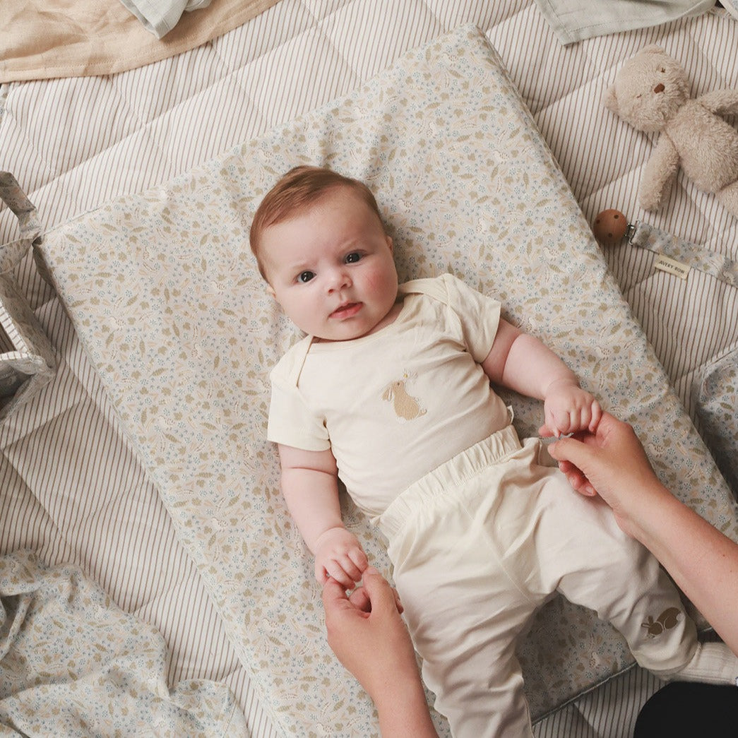 Baby lying on a changing table with a person's hand holding their foot, surrounded by toys and soft bedding.
