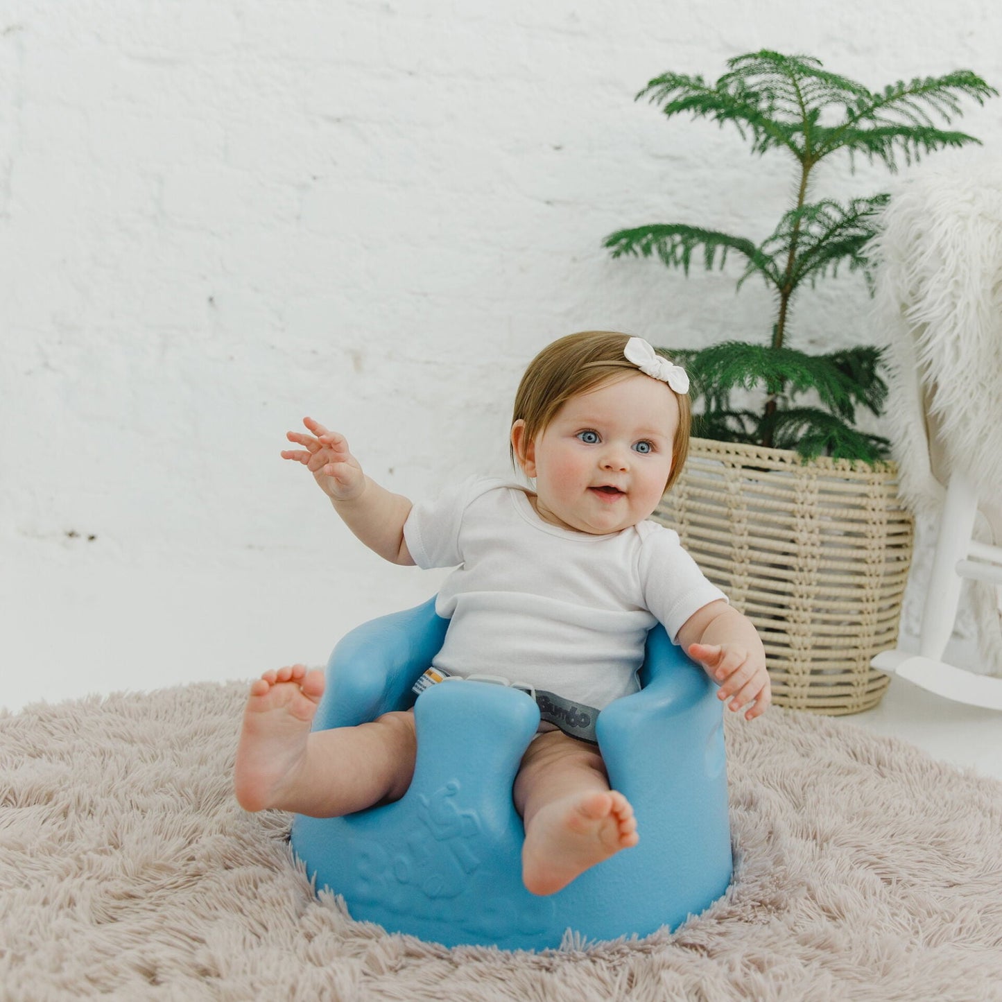 Baby sitting on a blue chair with a white wall and plant in the background