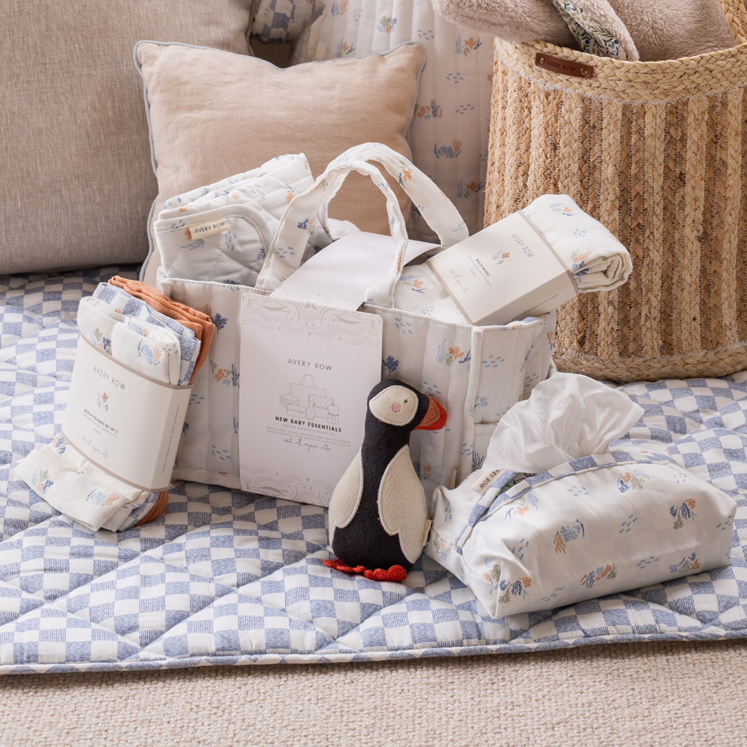 Baby gift set with diaper bag, blankets, and penguin toy on a checkered blanket.