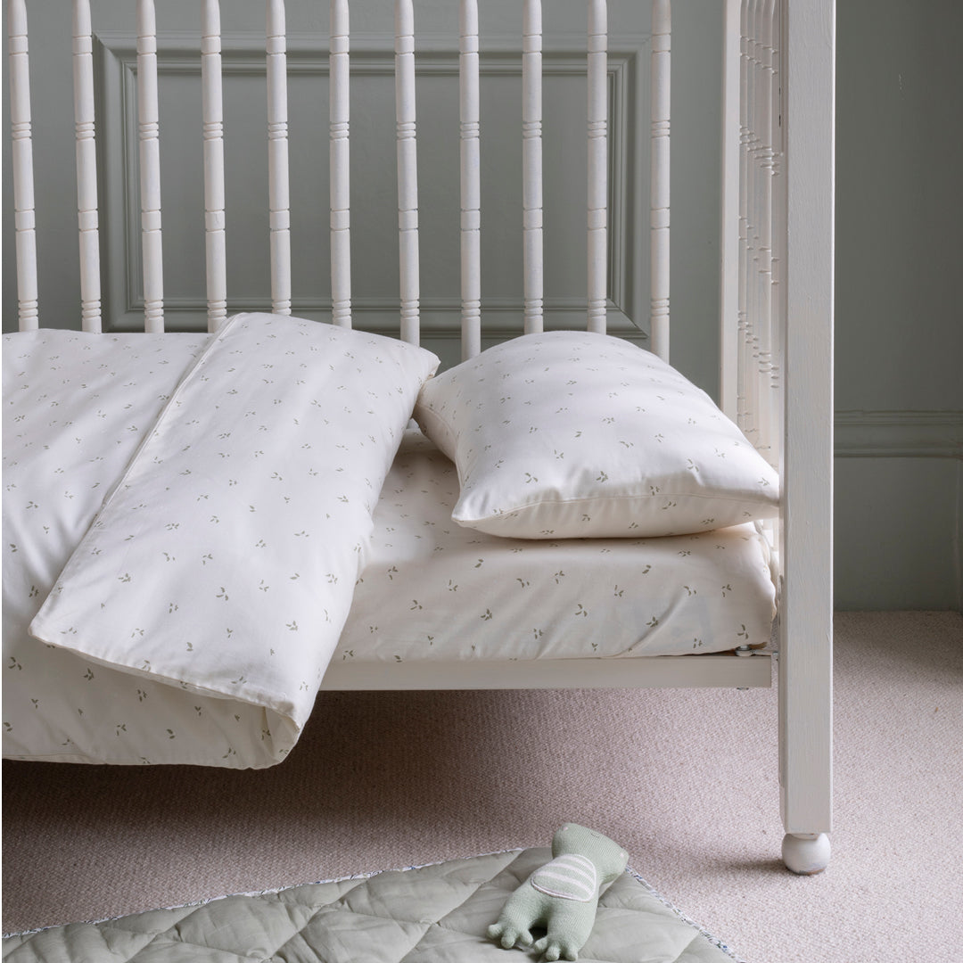 White crib with bedding and a toy on a carpeted floor against a gray wall.