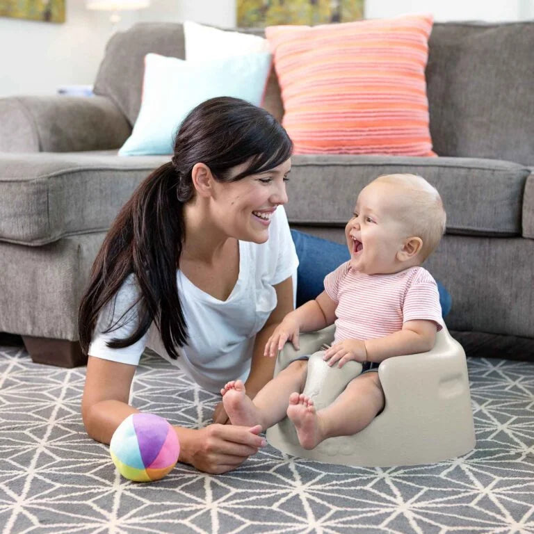 Woman and baby using a baby seat on a patterned rug with a couch in the background