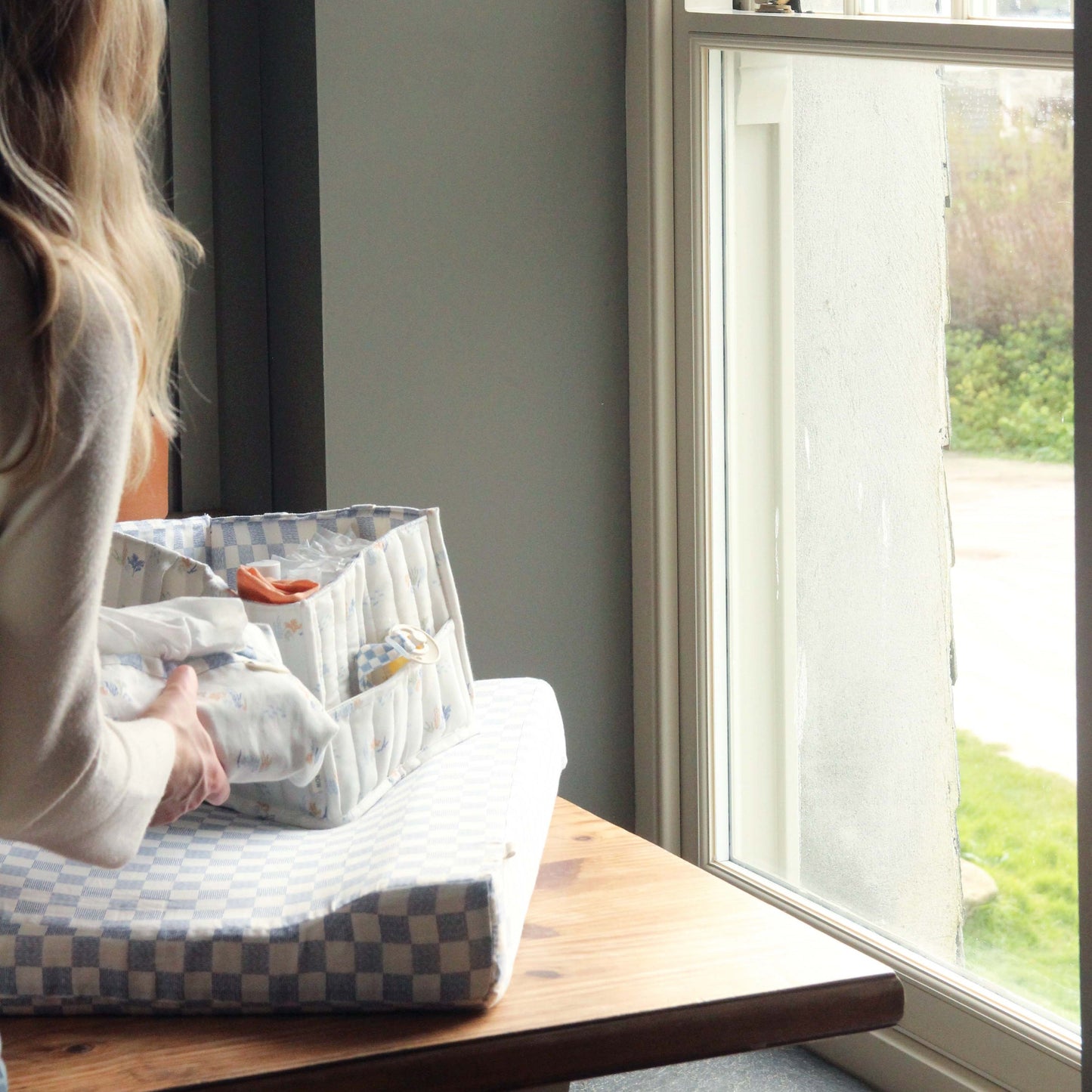 Person sitting by a window with a diaper bag and baby items on a wooden surface.