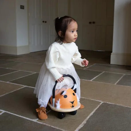Child in a white dress riding a toy car with a tiger design on a tiled floor.
