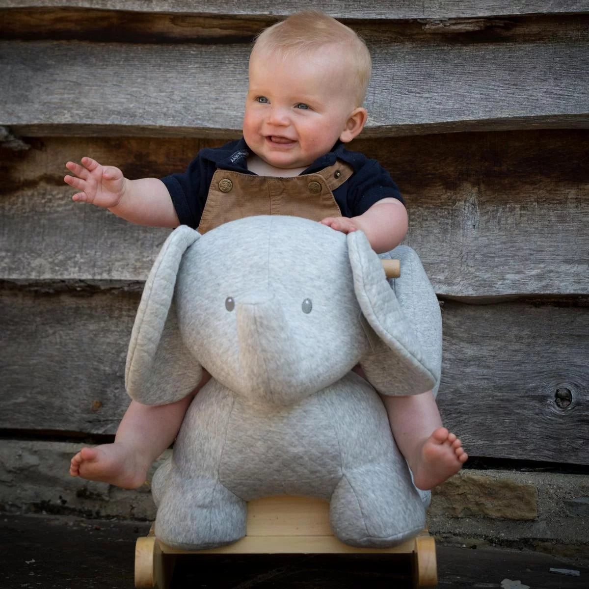 Baby sitting on a gray elephant toy with a wooden base against a wooden wall.