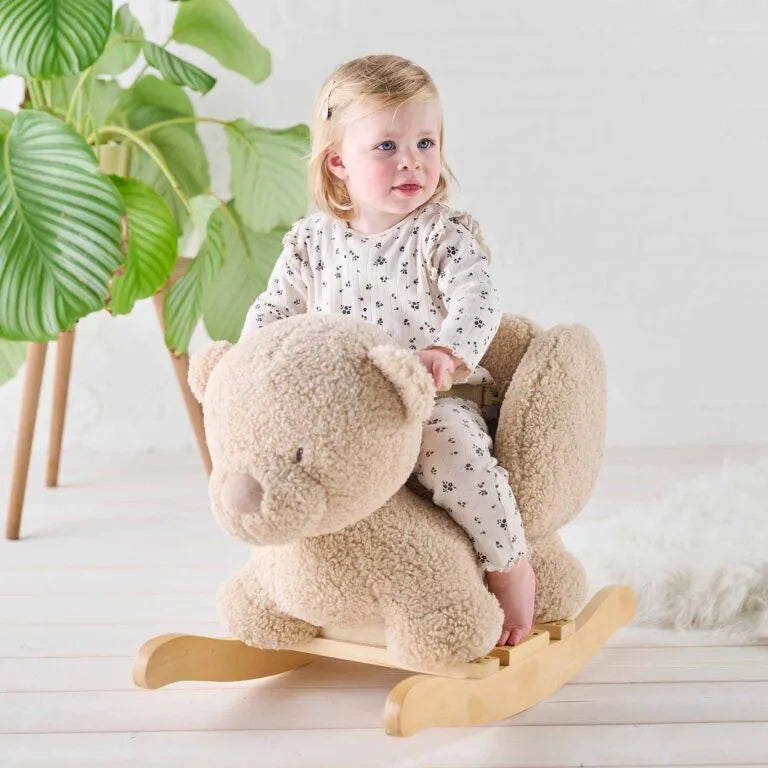 Child sitting on a teddy bear-shaped rocking toy in a bright room with plants.