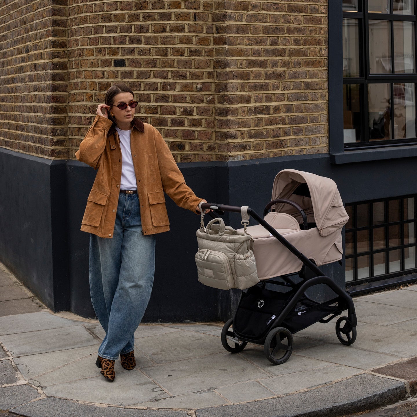 Woman in a brown jacket and jeans pushing a stroller against a brick wall.
