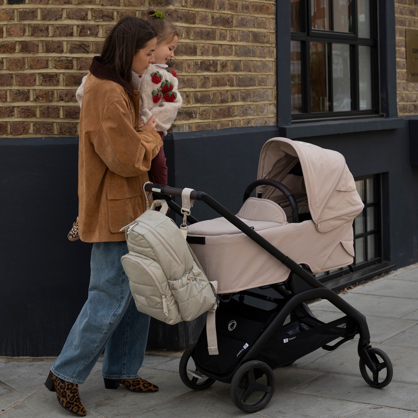 Woman pushing a beige stroller with a child holding a stuffed animal against a brick wall.