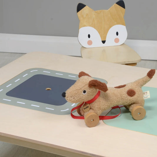 Plush toy dog with wheels on a table with a plush fox toy in the background.