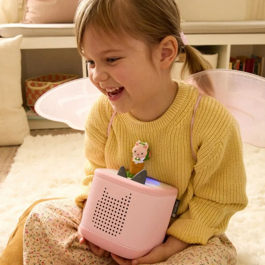 Child holding a pink toy with a screen, wearing a yellow sweater and fairy wings, in a cozy indoor setting.
