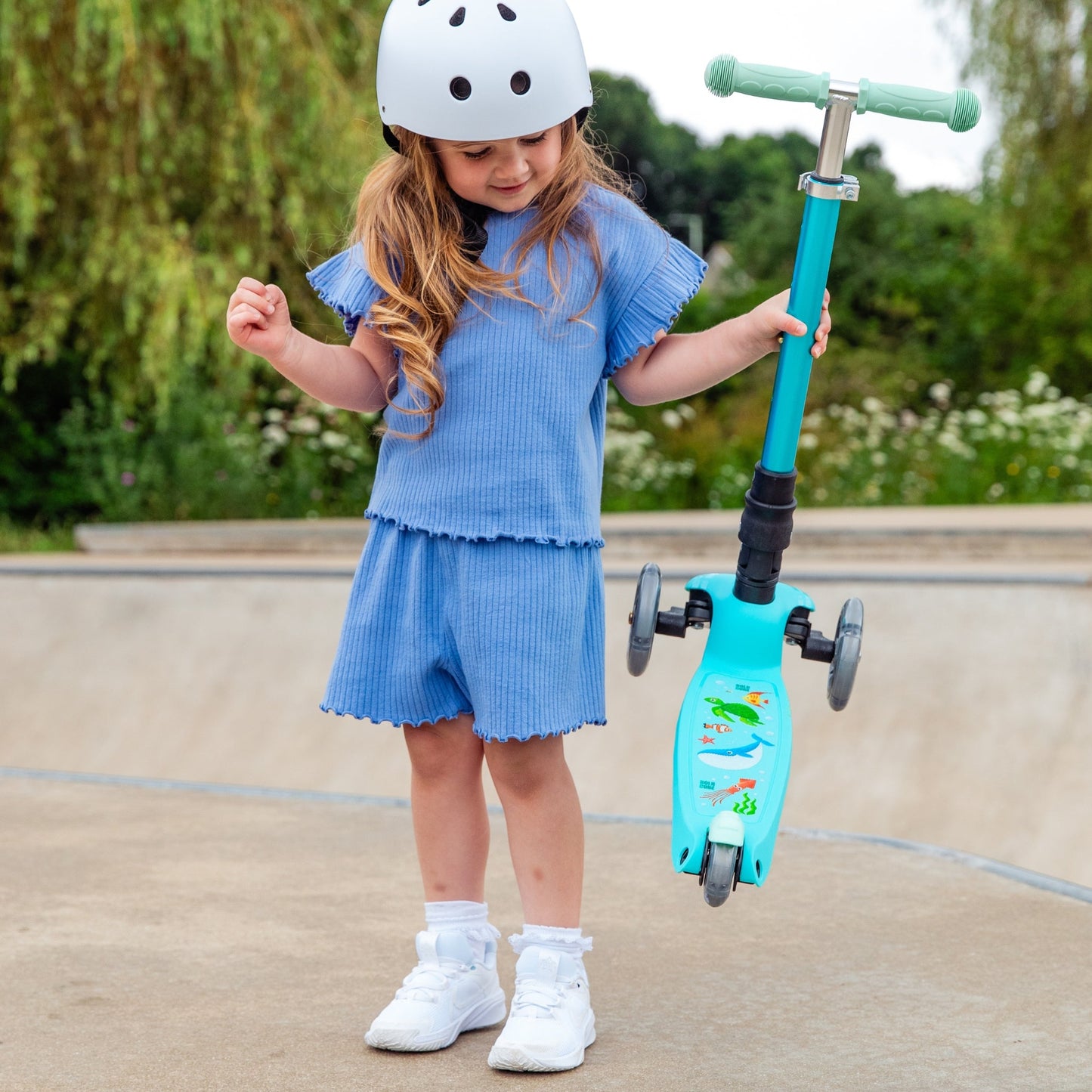 Child in a blue outfit with a helmet holding a turquoise scooter outdoors.