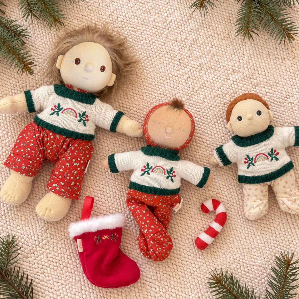 Three fabric dolls dressed in holiday sweaters and pants, arranged on a beige textured blanket with pine branches, a red stocking, and candy canes.