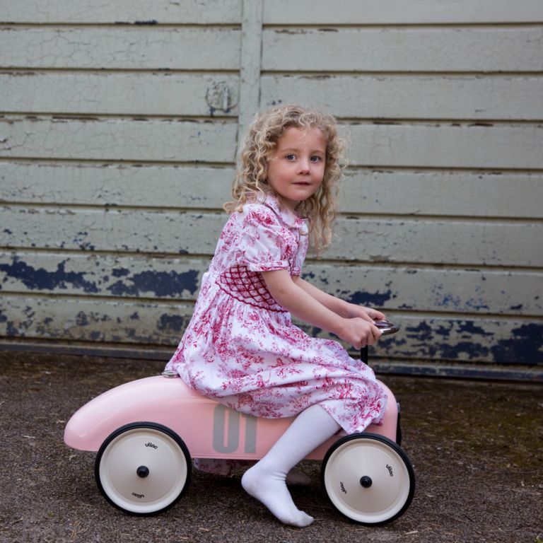 Young girl in a pink and white dress riding a pink toy car outdoors.