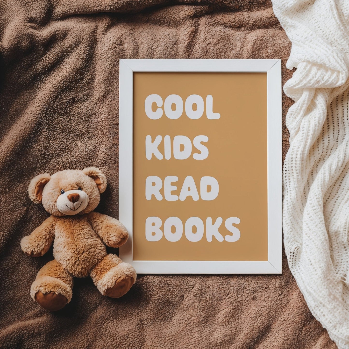 Teddy bear next to a framed sign with 'Cool kids read books' text on a brown blanket.