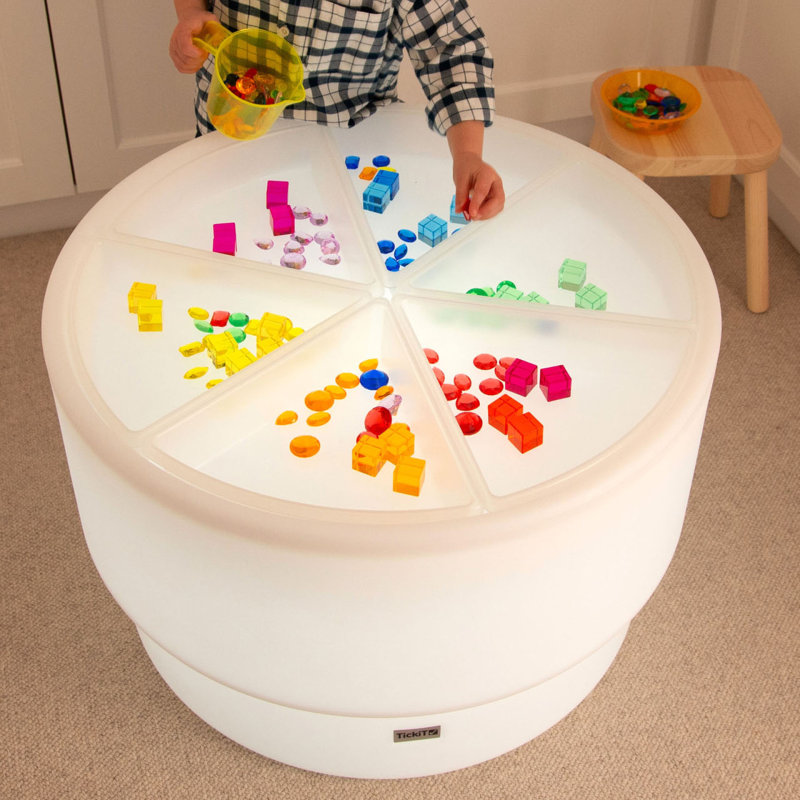 Children playing with colourful blocks sorted in plastic trays on a light-up table in a room.