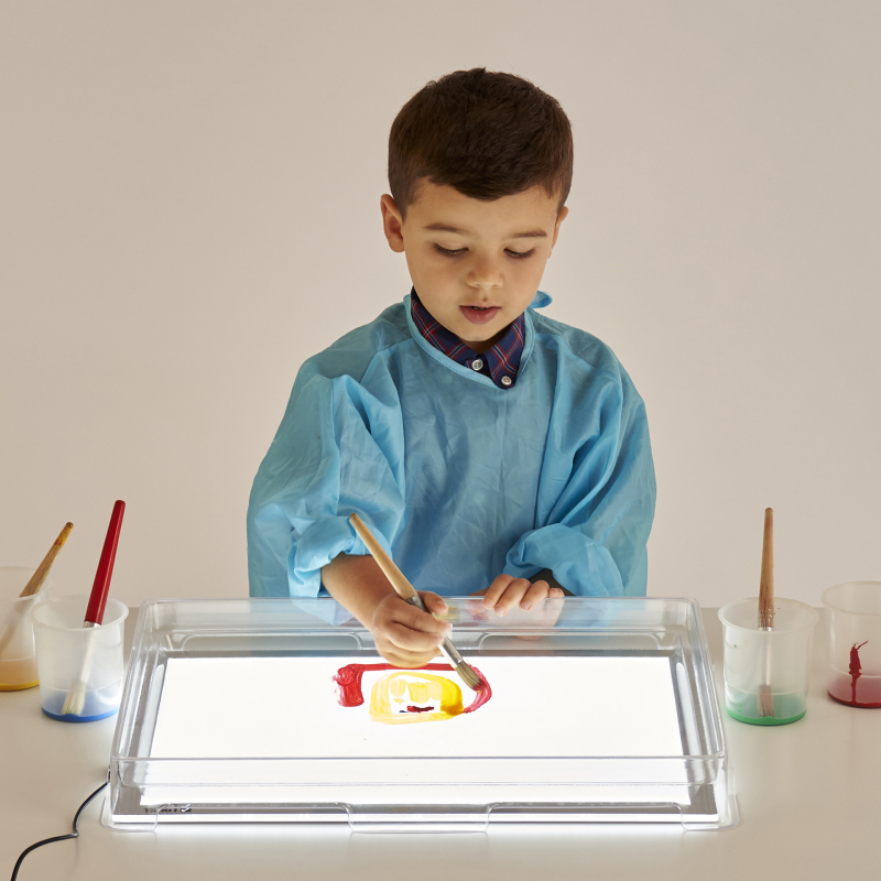 Child painting on a plasti cover on light panel with various art supplies around
