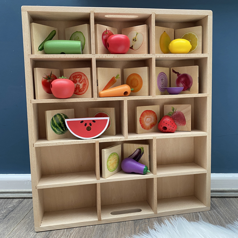 Wooden toy tray with colorful toy fruits and vegetables against a blue wall.