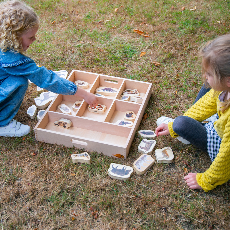 Two children playing with a wooden box containing animal stamps on grass