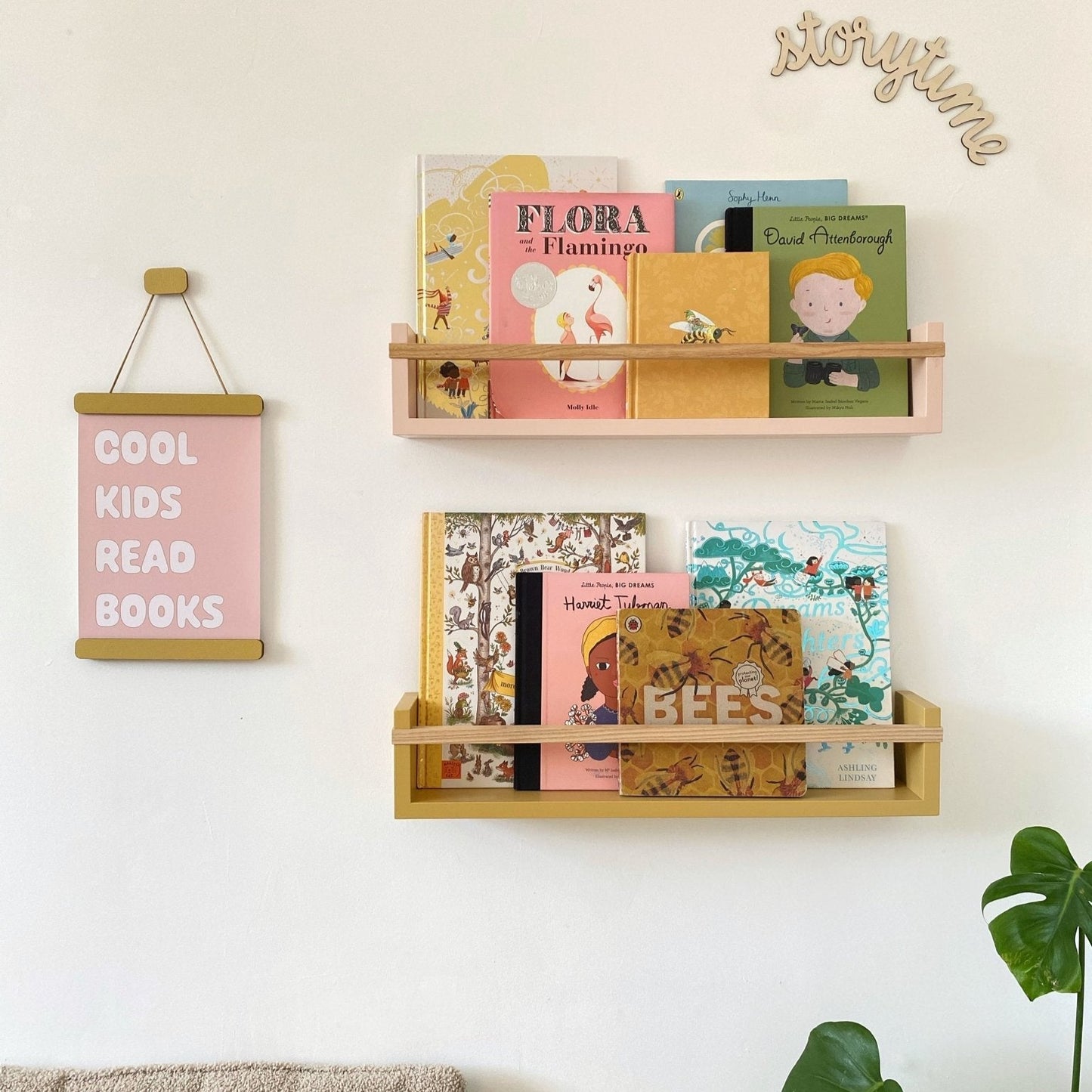 Wooden bookshelf with children's books on a wall, next to a pink sign and plant.