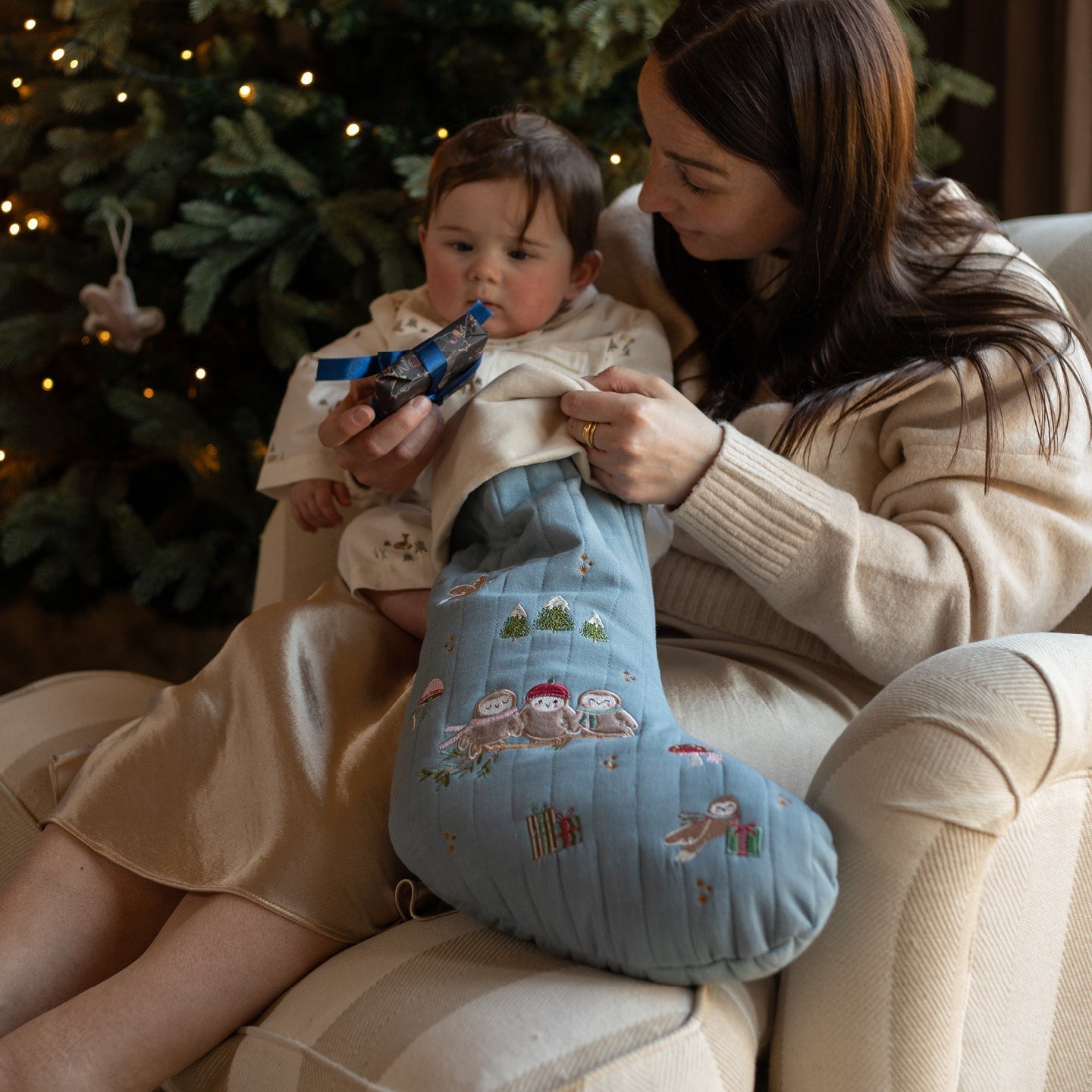 Woman holding a baby and a blue embroidered stocking in front of a decorated Christmas tree.
