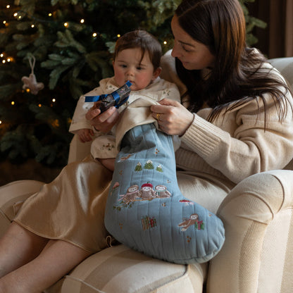 Woman holding a baby and a blue embroidered stocking in front of a decorated Christmas tree.