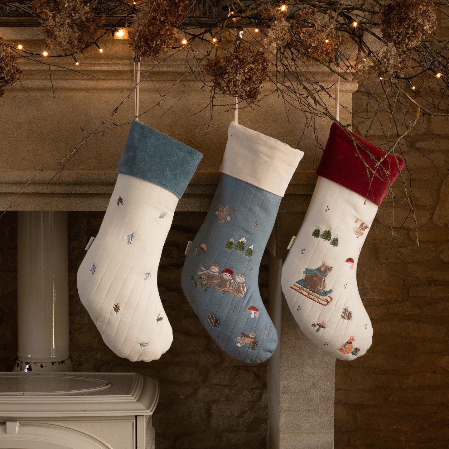 Three Christmas stockings hanging on a fireplace mantle with decorative lights and branches.
