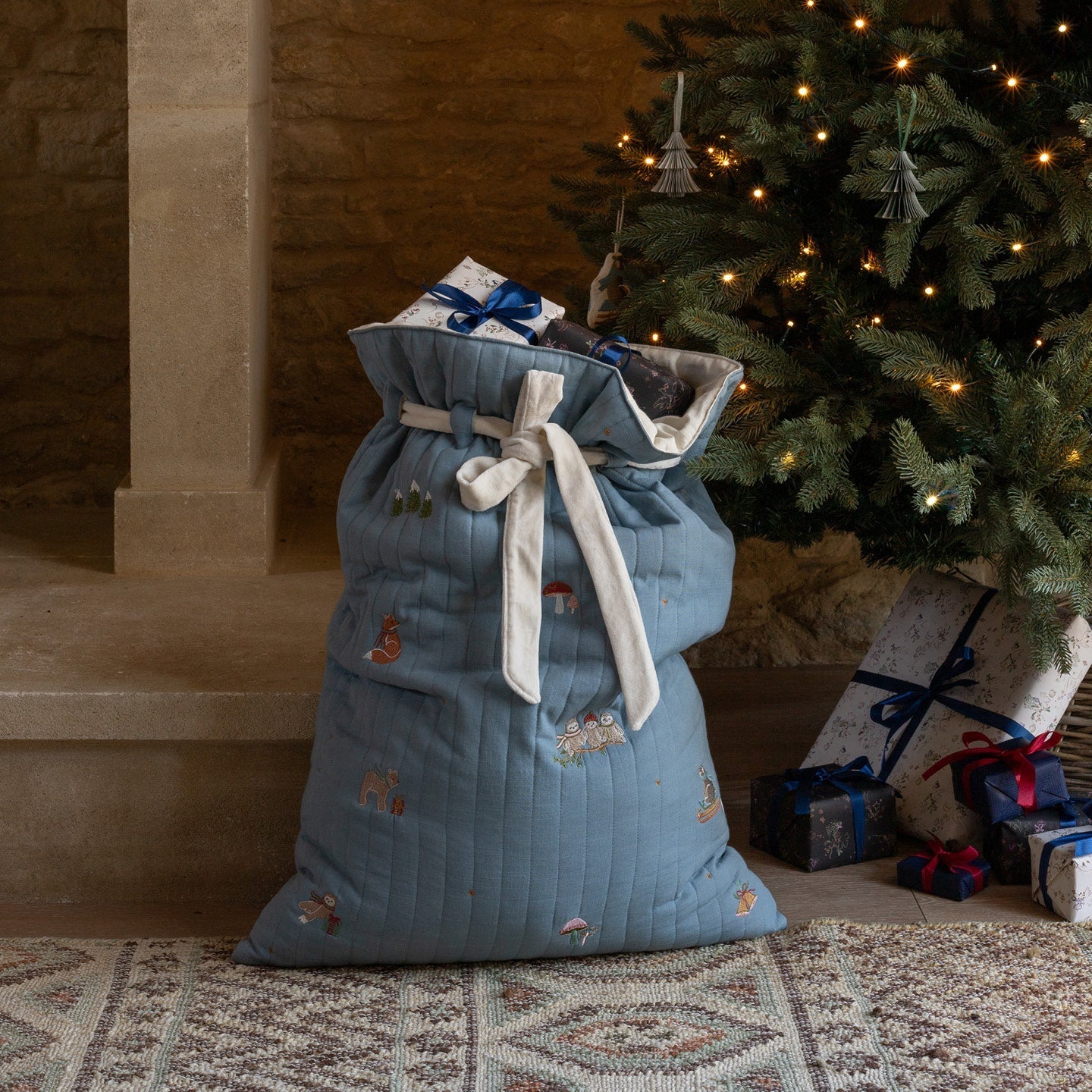 Blue gift sack with embroidered designs in front of a decorated Christmas tree.