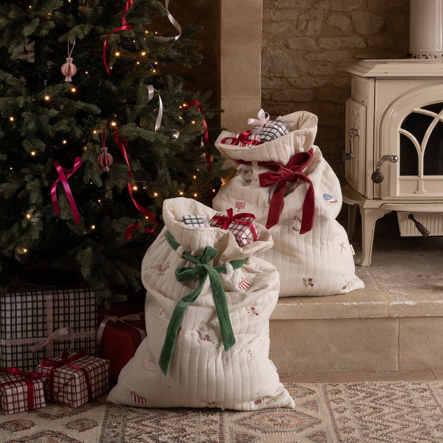 Two white gift sacks with red and green ribbons in front of a decorated Christmas tree and stone fireplace.