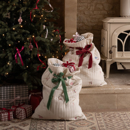 Two white gift sacks with red and green ribbons in front of a decorated Christmas tree and stone fireplace.