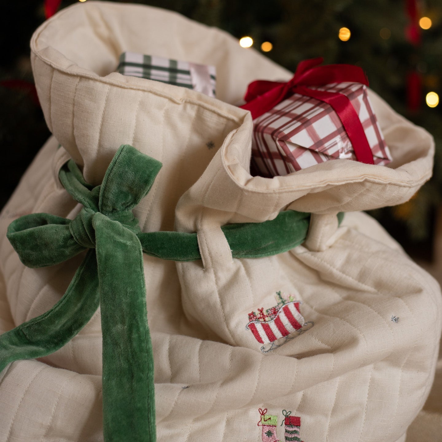Beige fabric bag with green velvet tie, embroidered details, and small wrapped gifts inside against a blurred festive background.