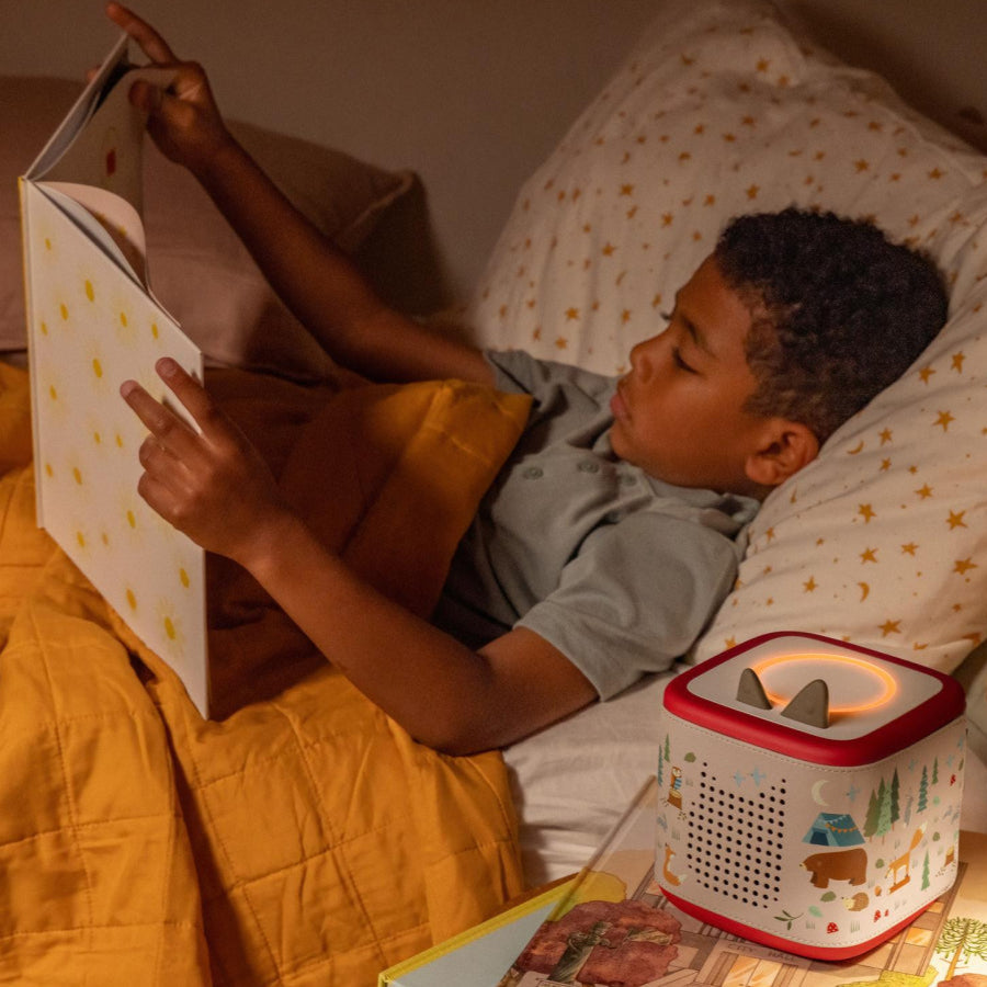 Child reading a book in bed with a night light on a wooden nightstand.