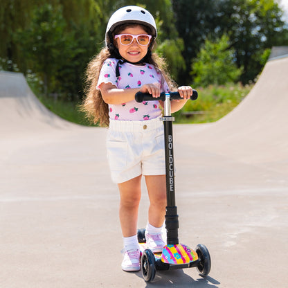 Child riding a scooter with a colorful deck on a paved path with greenery in the background
