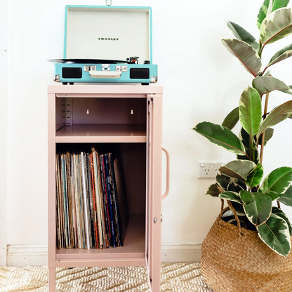 Record player on a pink cabinet with vinyl records, a plant, and colorful abstract art in the background.
