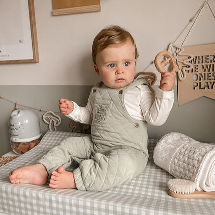 Baby sitting on a bed with checkered sheets, surrounded by wooden toys and decorations.