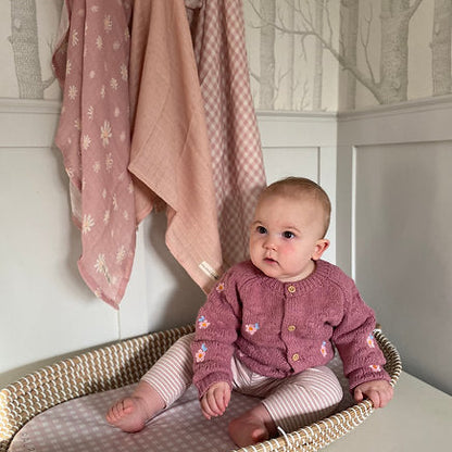 Baby in a pink cardigan sitting in a crib with pink blankets in the background
