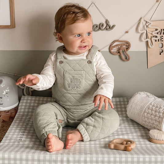 Baby in a quilted overalls sitting on a checkered changing table with toys around.