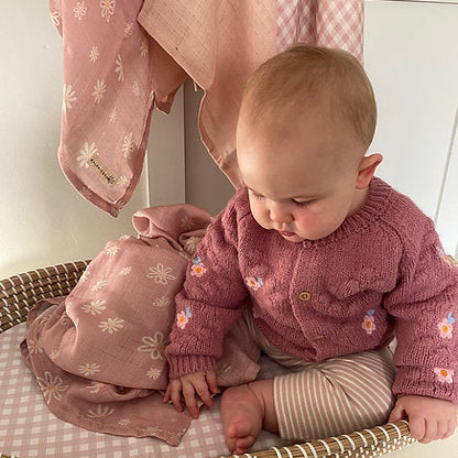 Baby in pink outfit sitting on a woven basket with pink clothing in the background