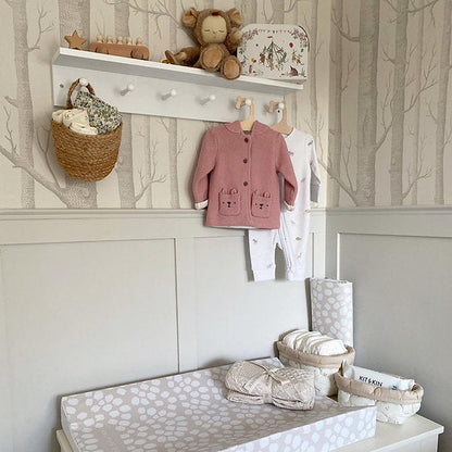 Nursery setup with changing table, baby clothes, and toys against a tree-patterned wall.