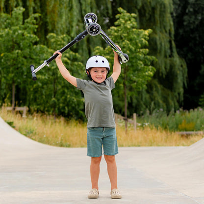 Child holding a scooter above their head with a helmet on, standing on a paved path with greenery in the background.