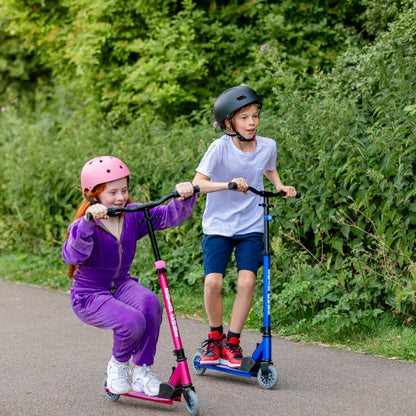 Two children riding scooters on a path with greenery in the background