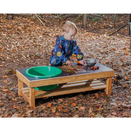 Child playing at a wooden outdoor table with green and silver bowls on a leaf-covered ground.