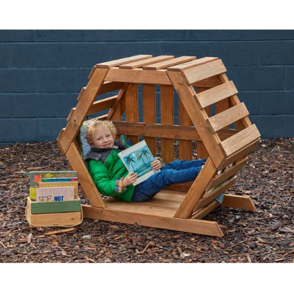 Child sitting in a wooden playhouse reading a book with books nearby on a mulched ground.