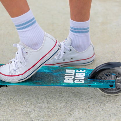 Person wearing white sneakers with red accents on a teal scooter with 'Bold Cube' branding.