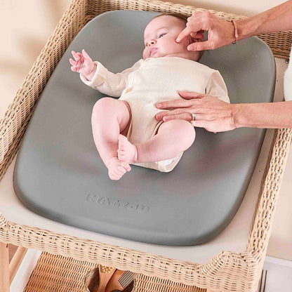 Baby sitting on a gray changing pad with a person's hands near, on a wicker chair.
